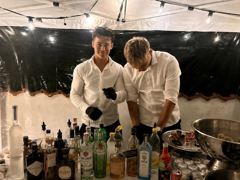 To bartendere i hvide skjorter og sorte handsker der laver cocktails ved en indendørs event med spiritus og flasker på bardisken